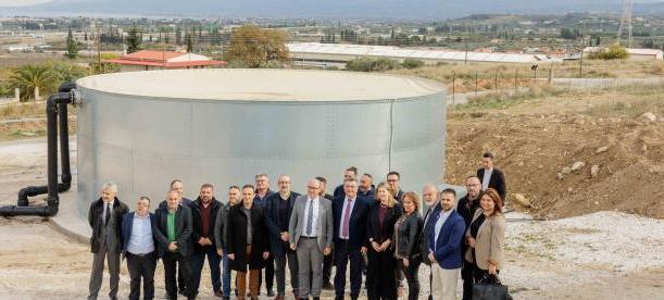 A group of people posing in front of a circular water tank in an outdoor setting, with a rural landscape and distant mountains in the background. The individuals are dressed in professional attire.